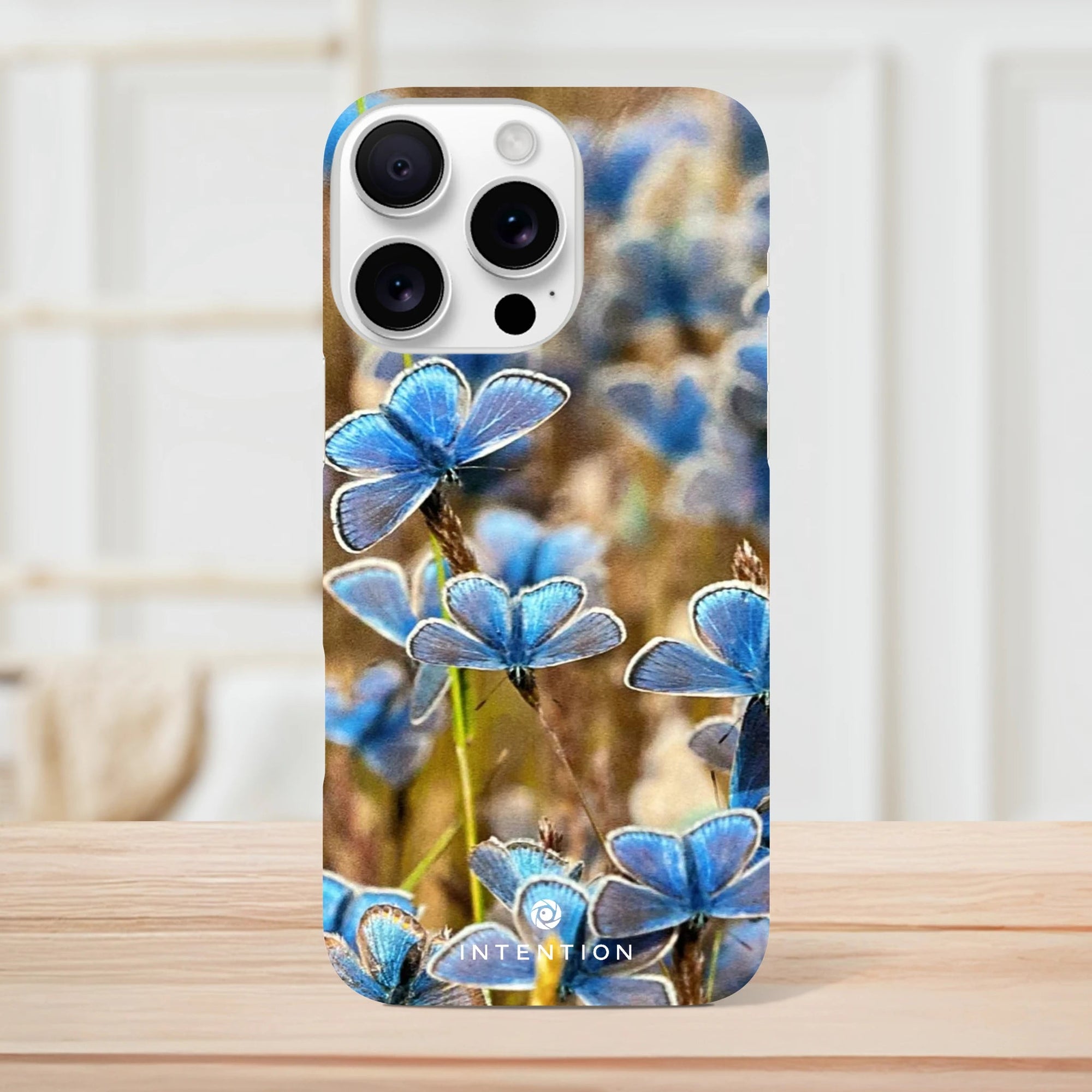 A smartphone case with a photo art design featuring multiple blue butterflies on grass, displayed upright on a wooden surface with a light-colored, blurred background. Textures and the word INTENTION appear near the bottom of the case.
