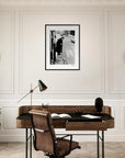 A modern office with mindful home decor features a brown leather chair, dark desk, desk lamp, potted plant, decorative bust, and brass telescope. A black-and-white photo in a gallery-quality frame hangs on the white paneled wall above the desk.