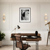 A modern office with mindful home decor features a brown leather chair, dark desk, desk lamp, potted plant, decorative bust, and brass telescope. A black-and-white photo in a gallery-quality frame hangs on the white paneled wall above the desk.