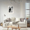 A bright living room with a white sofa, neutral-toned pillows, a floor lamp, and a small wooden table. A fine art print in a light frame hangs above the sofa, adding mindful home decor to the space. White curtains cover the window on the right.