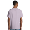 A person is standing facing away from the camera, wearing a light purple short-sleeve t-shirt and light blue jeans. This mindful apparel features a small black logo printed below the neckline on the back.