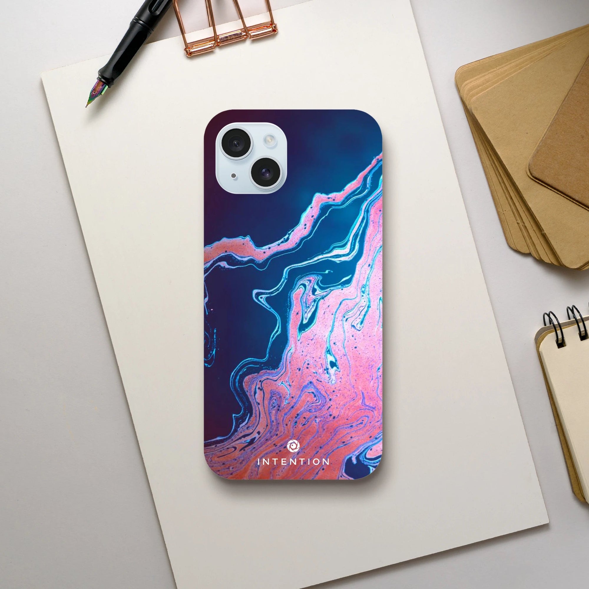 A smartphone with a blue and pink abstract swirl case labeled INTENTION lies on a blank sheet of paper, surrounded by a pen, brown folders, and a small spiral notebook—capturing photo art textures in an everyday setting.