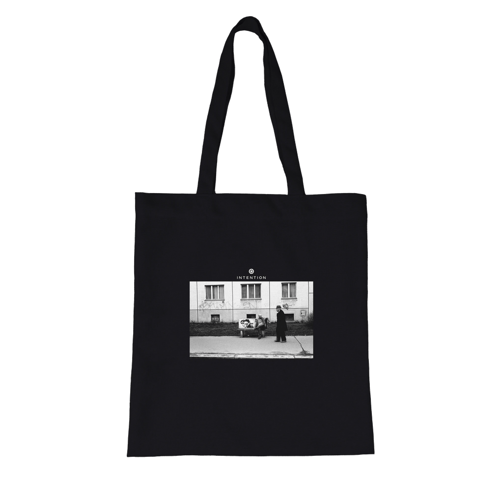 This black tote bag with long handles features an art print—a black and white photo of two people on a sidewalk in front of a building—with "INTENTION" printed above, crafted with conscious fashion in mind.