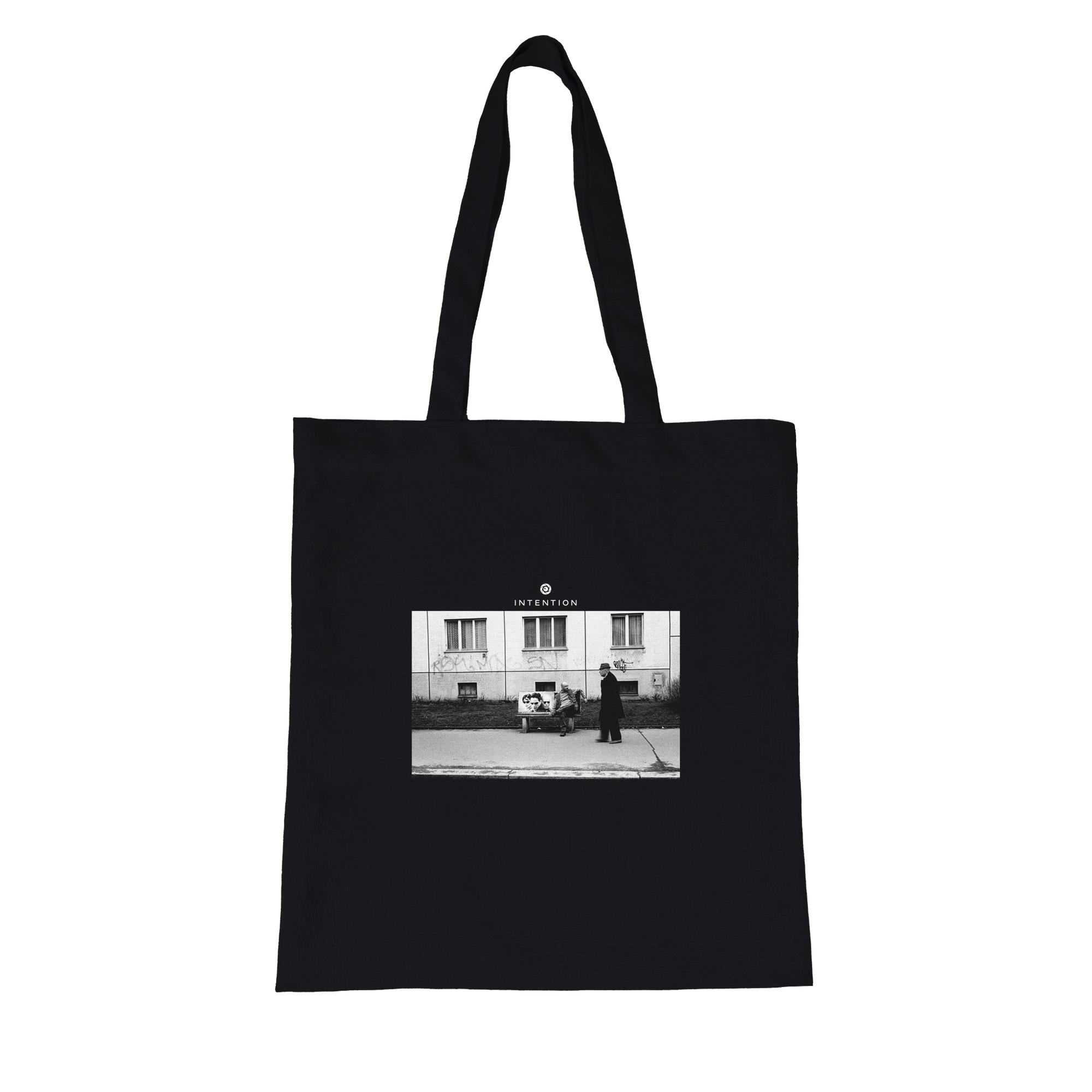 This black tote bag with long handles features an art print—a black and white photo of two people on a sidewalk in front of a building—with "INTENTION" printed above, crafted with conscious fashion in mind.