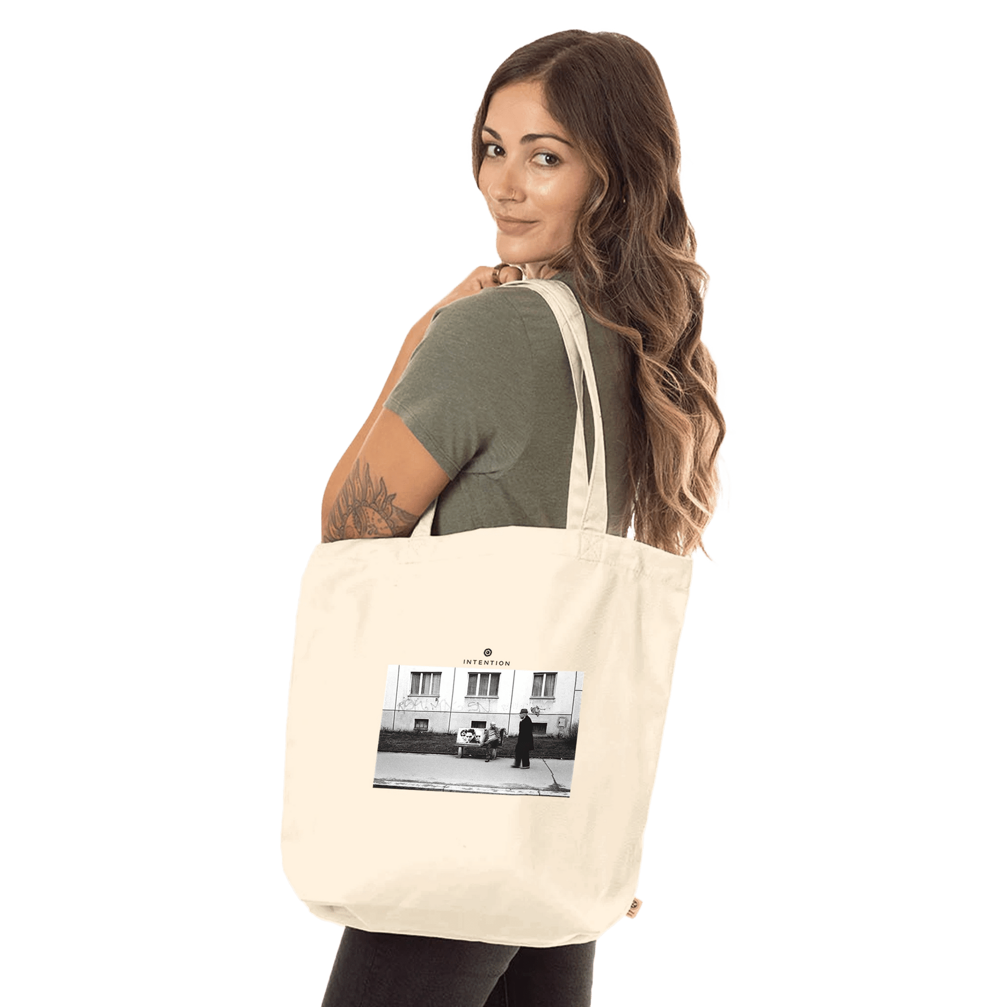 A woman with long brown hair, wearing a green t-shirt, holds a cream-colored tote bag over her shoulder. This piece of conscious fashion features an art print of a black-and-white street photo. The background is plain white.