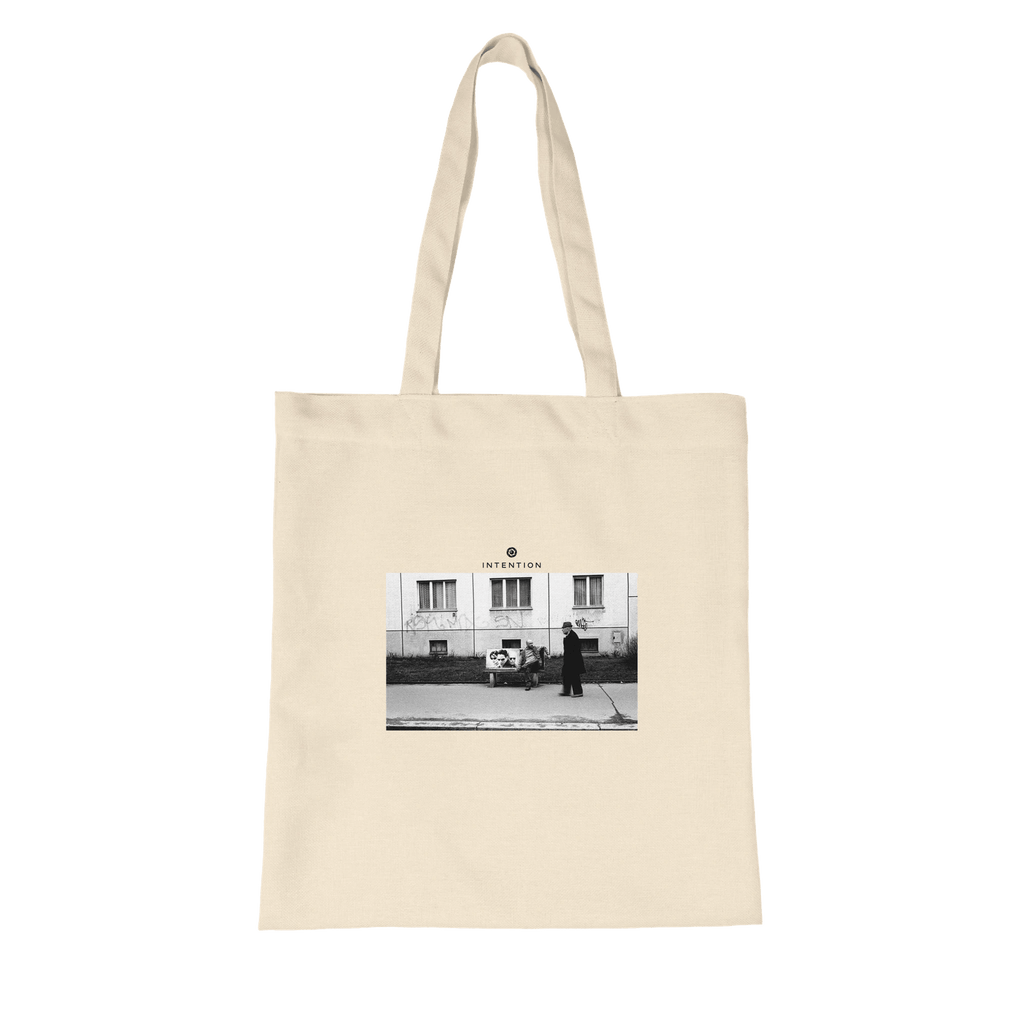 A natural tote bag made from organic material, featuring a black and white art print of a person walking past another sitting outside a building, with the word INTENTION printed above the image.